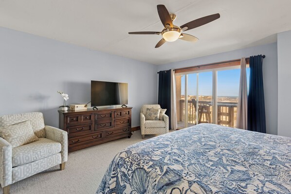 A bedroom with a bed, a dresser, a TV, two chairs, a ceiling fan, and a large window with curtains. - Sea Mist: Oceanfront | Pool and Tennis Access (Nags Head)
