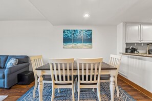 A dining area with a table and chairs, a painting on the wall, a kitchen area, and a gray sofa.