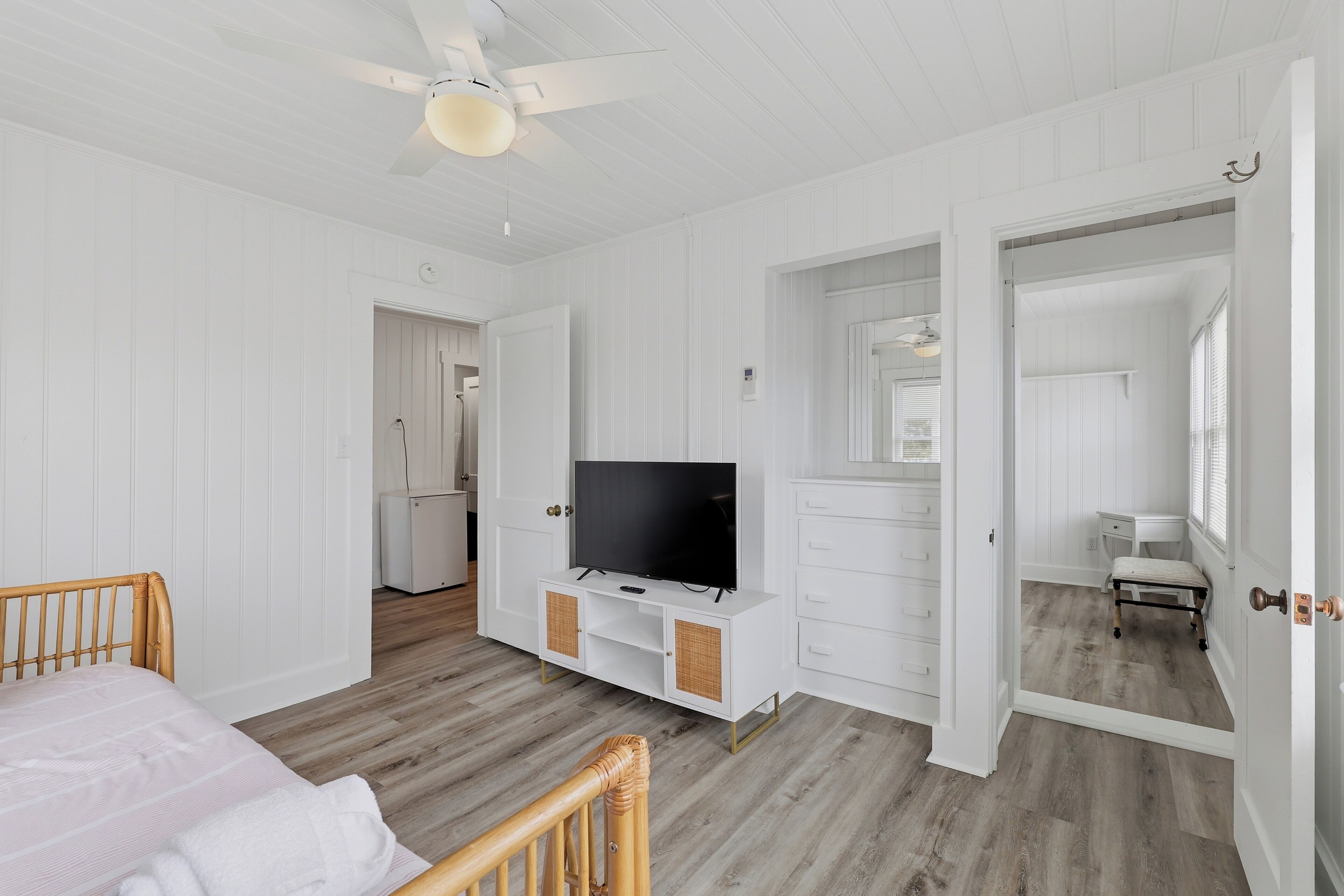 A bedroom with a bed, a TV on a white cabinet, a ceiling fan, and a door leading to another room.