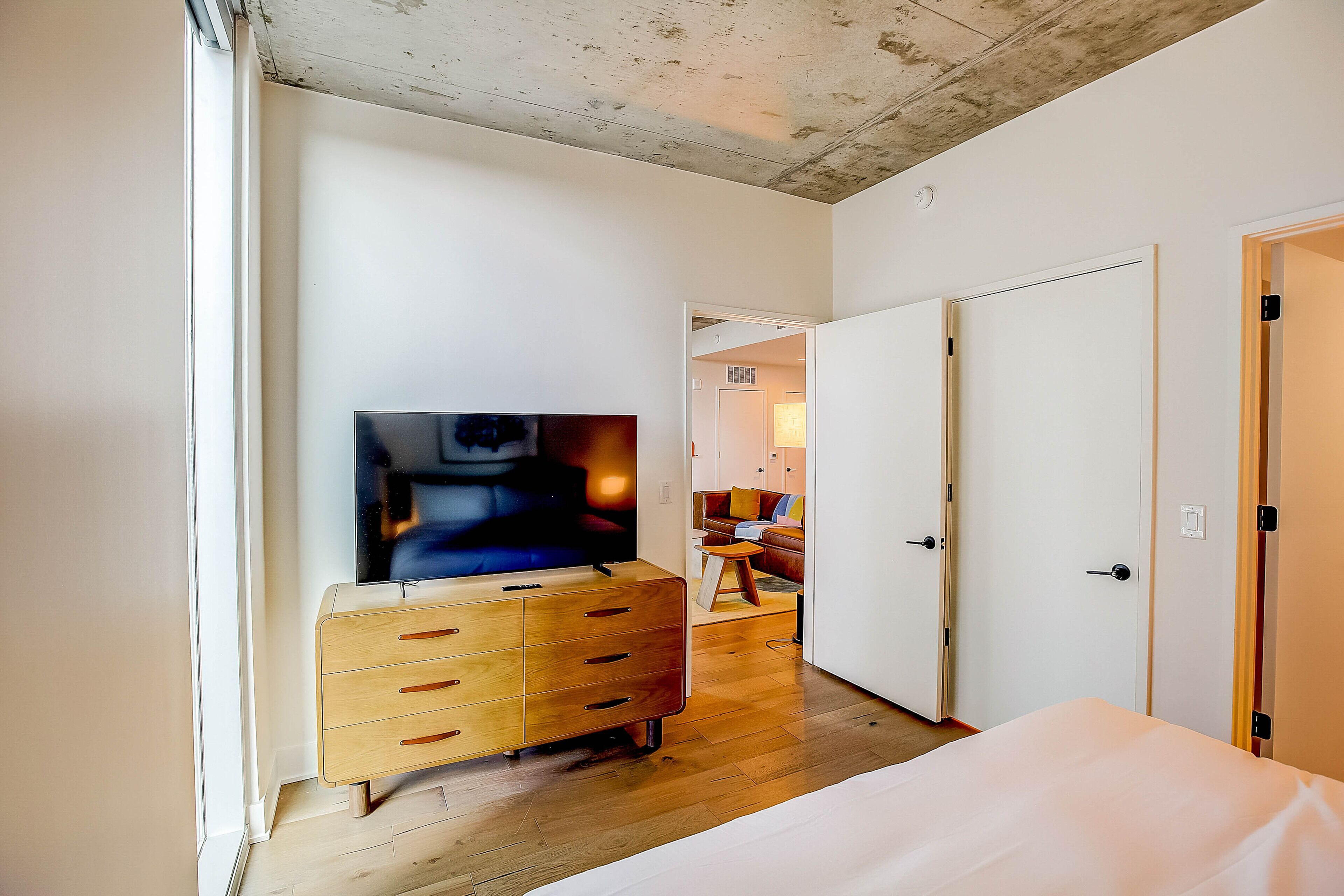 Une chambre à coucher comprenant une commode en bois, un téléviseur à écran plat, un lit et un couloir menant à un espace de vie.