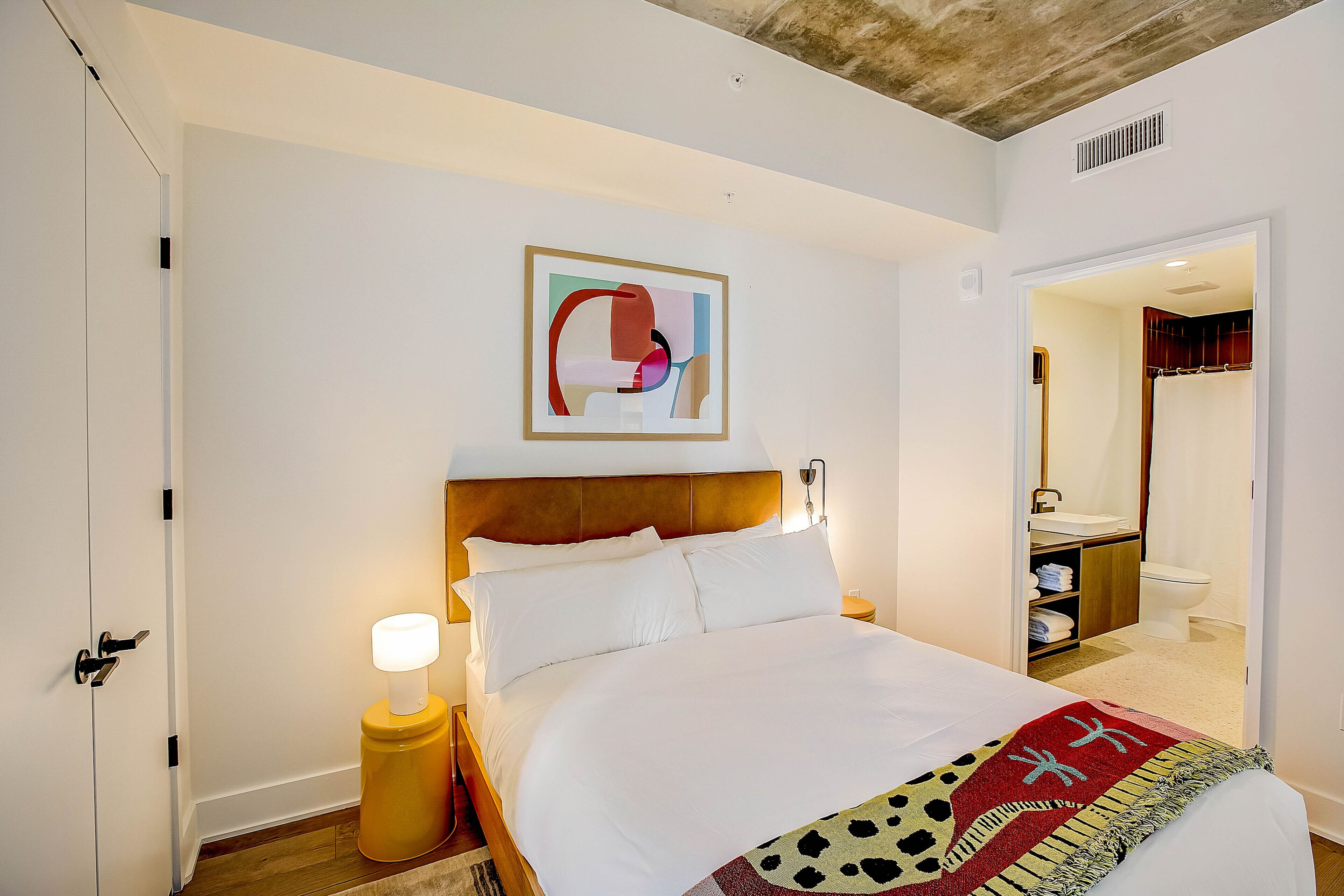 A hotel room with a large bed, a bedside lamp, a framed abstract artwork, and a view into the bathroom.