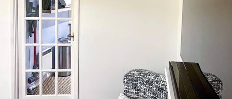Una habitación con un piano, un reposapiés marrón, un tapete estampado y una puerta blanca que da a un área de almacenamiento.