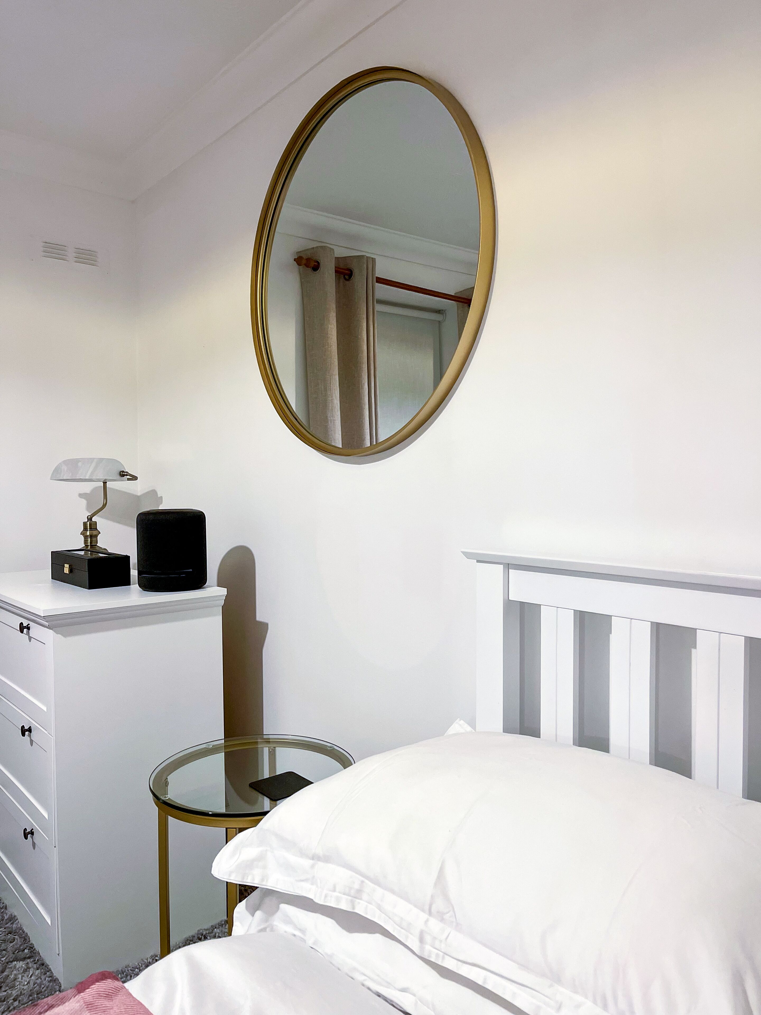 A bedroom with a white dresser, a round mirror, a bedside table, and a bed with white bedding.