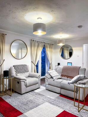 A modern living room with two sofas, a coffee table, and a TV. There are curtains, a mirror, and a ceiling light fixture.