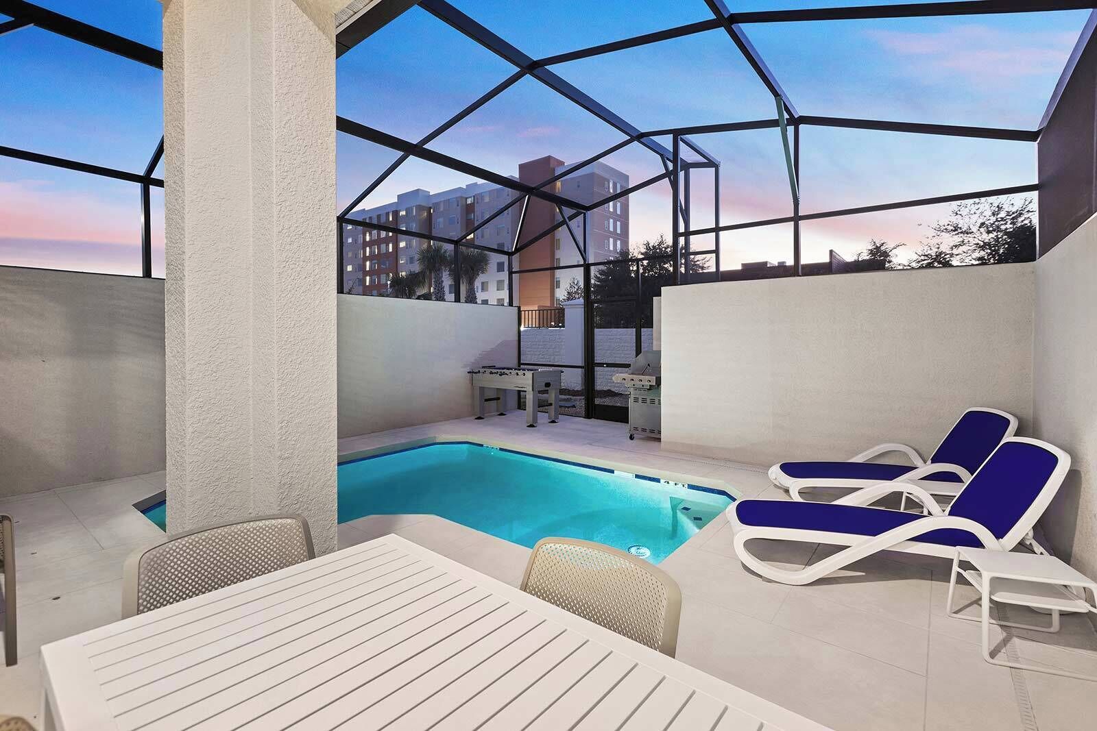 A covered outdoor pool area with lounge chairs, a table, and a grill.