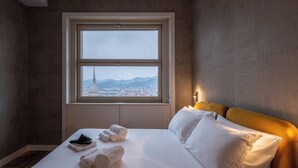 A hotel room with a large window, a bed with white linens, and a view of a cityscape and mountains. - Appartamento Luxury con Balcone (Torino)