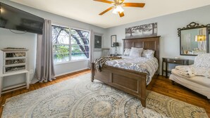 Une chambre à coucher avec un lit en bois, un ventilateur de plafond, une grande fenêtre avec des rideaux, une table de chevet, une commode et un miroir.
