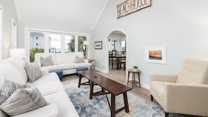 Un salon spacieux avec un canapé blanc, un fauteuil beige et une table basse en bois. On aperçoit à l’arrière-plan un coin repas agrémenté d’un lustre.