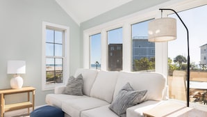 Un salon moderne avec un canapé blanc, un pouf bleu et une table d’appoint en bois sur laquelle se trouve une lampe. De grandes fenêtres offrent une vue sur la plage et les bâtiments.
