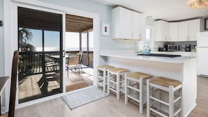 Une cuisine moderne avec des armoires blanches, un coin bar avec des tabourets et une vue sur l’océan grâce à une porte-fenêtre coulissante.