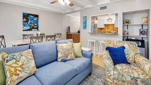A living room with a blue sofa, matching armchairs, and a ceiling fan. There is a dining area with a table and chairs, and a kitchen with a tiled backsplash. - Steps to East Beach & Pet Friendly w/ 2 King Beds (St. Simons Island)