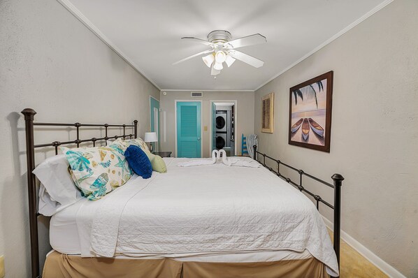 A bedroom with a bed, a ceiling fan, a door, a wall-mounted picture, and a laundry area. - Steps to East Beach & Pet Friendly w/ 2 King Beds (St. Simons Island)