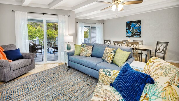 A living room with a sofa, a chair, a dining table with chairs, a ceiling fan, and a view through glass doors to an outdoor patio. - Steps to East Beach & Pet Friendly w/ 2 King Beds (St. Simons Island)