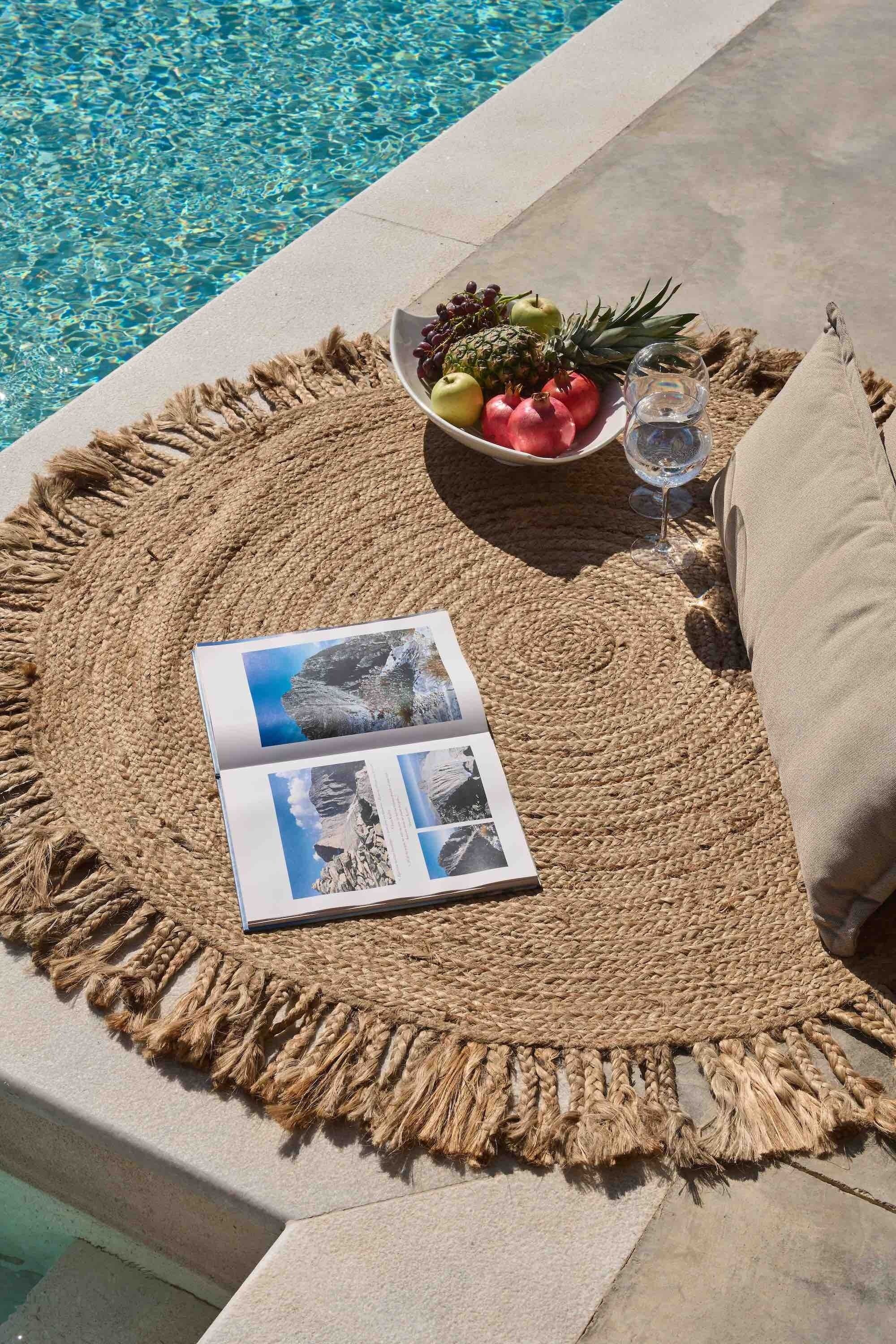 A poolside area with a woven mat, a bowl of fruit, a glass of water, and a magazine.