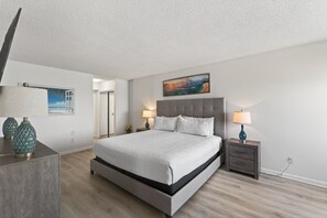 A modern bedroom with a large bed, bedside tables, a nightstand, a painting, and a door leading to another room.