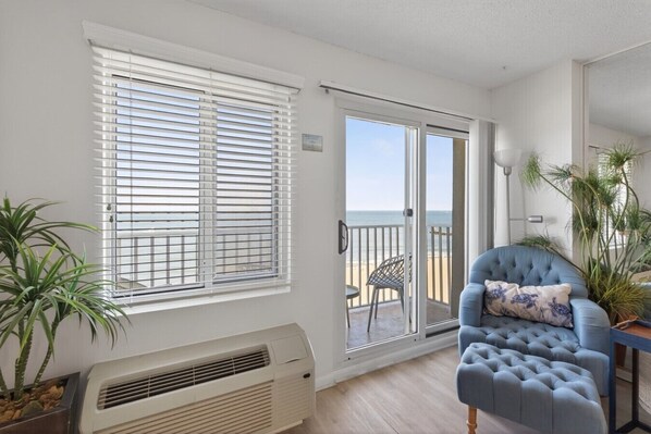 A room with a large window, a sliding glass door leading to a balcony, a blue armchair, and a potted plant.