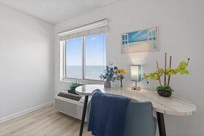 A room with a large window, a table with a vase of flowers, a lamp, and a painting of a beach scene.