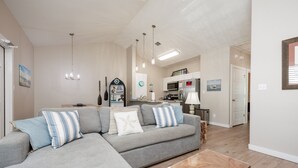 Un salon moderne avec un canapé d’angle gris, une table basse en bois, des luminaires suspendus et une cuisine en arrière-plan.