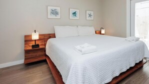 Un lit bien fait, avec une literie blanche, une tête de lit en bois et une table de chevet sur laquelle se trouve une lampe. Trois tableaux encadrés sont accrochés au mur au-dessus du lit.