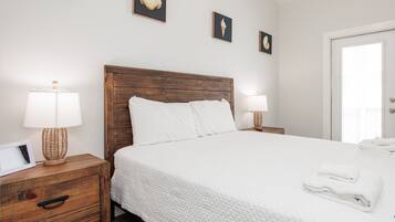 Un dormitorio limpio y bien iluminado con cabecera de madera, ropa de cama blanca y mesita de noche con lámpara.