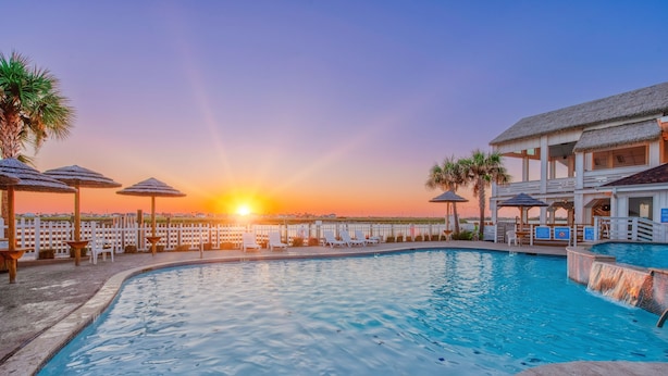 Ein Pool mit Liegestühlen und Sonnenschirmen, ein Gebäude mit einem Strohdach und ein Sonnenuntergang im Hintergrund.