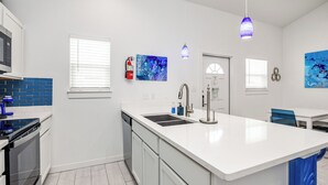 A modern kitchen with a central island, white cabinetry, a built-in oven, and a dishwasher. There are two pendant lights, a fire extinguisher, and artwork on the walls. - Experience a ‘blu-tiful’ retreat: Pet-Friendly, Private Patio, Pool & More (Corpus Christi)
