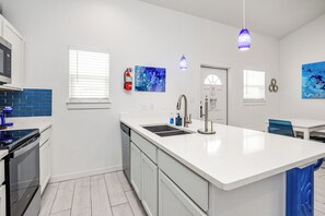 A modern kitchen with a central island, white cabinetry, a built-in oven, and a dishwasher. There are two pendant lights, a fire extinguisher, and artwork on the walls.
