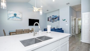 A modern kitchen with a large island, white cabinetry, and a ceiling fan. There are wall-mounted artworks and a dining area with wooden chairs and a table. - Spacious Coastal Retreat | Heated Pools, Slides & Resort Amenities (Corpus Christi)