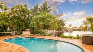 Une piscine en forme de courbe, dotée d’une terrasse carrelée, entourée de palmiers et offrant une vue sur une étendue d’eau.