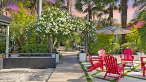 Een tropische tuinpatio met rode Adirondack-stoelen, een pad en een pergola met witte bloemen.