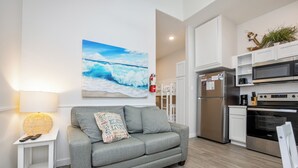 Une cuisine moderne avec des appareils en acier inoxydable, un canapé gris agrémenté d’un coussin sur le thème de la plage, et un grand tableau représentant une vague océanique accroché au mur.