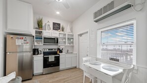 Une cuisine moderne avec des meubles blancs, un réfrigérateur en acier inoxydable, un four à micro-ondes et un four traditionnel. On y trouve une table à manger avec des chaises blanches et une fenêtre équipée de stores.