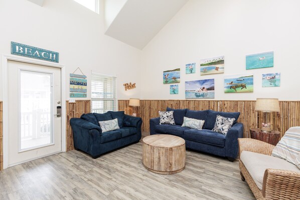 Ein Zimmer mit zwei blauen Sofas, einem runden Holztisch und einem Korbstuhl. An den Wänden befinden sich Bilder von Strandmotiven.