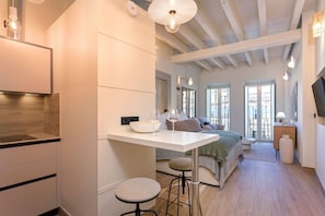 A modern kitchen with a white countertop, bar stools, and a dining area with a bed and a window.