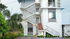 A modern multi-story building with a central staircase, surrounded by a landscaped area with palm trees and a patterned walkway. - Diamond Residency (Ambalapuzha)