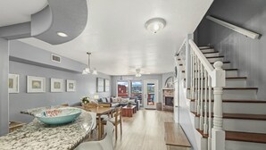 Una cocina moderna con encimera de granito, un área de comedor con mesa y sillas, una escalera con barandilla blanca y vistas al exterior a través de un ventanal.