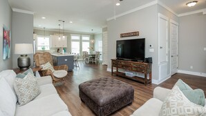 A spacious living room with a large television, comfortable seating, a ceiling fan, and a kitchen in the background.