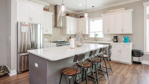 A modern kitchen with white cabinets, a stainless steel refrigerator, a large island with bar stools, and a stone backsplash.