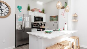A modern kitchen with a central island, stainless steel appliances, wicker stools, and decorative wall art. - Pirate Ship Playground | Tropical Pool & Family Fun (Corpus Christi)