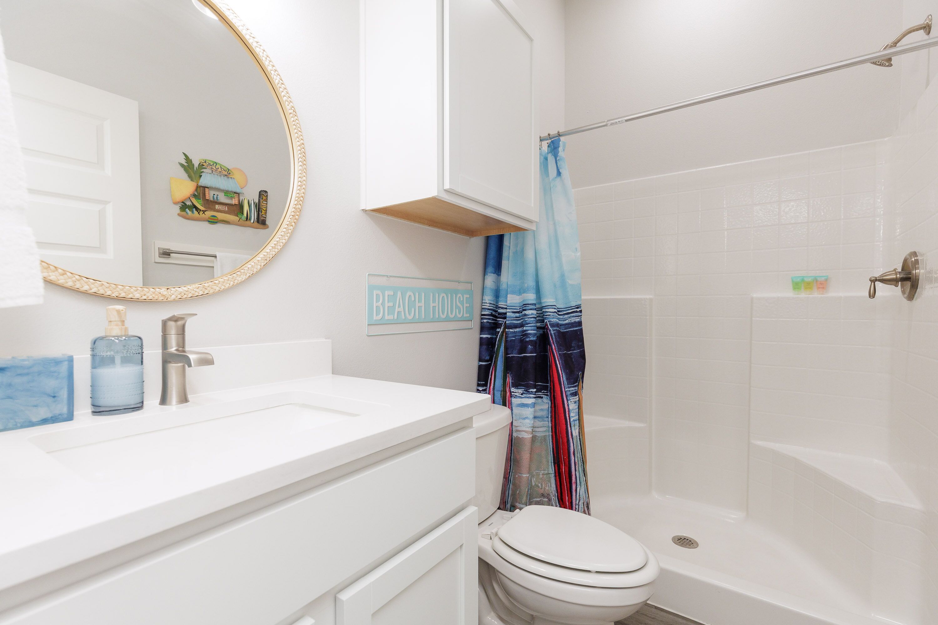 Et badeværelse med en hvid vask, toilet og brusebad med blå gardin, samt et skilt der står 'BEACH HOUSE'.
