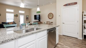 A kitchen with a granite countertop, stainless steel sink, and dishwasher. There is a ceiling fan, a TV mounted on the wall, and a door labeled "Beach House". - Pet-Friendly Beach Escape | Two Heated Pools & Coastal Charm (Corpus Christi)