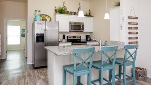 A kitchen with white cabinets, a stainless steel refrigerator, a microwave, a coffee maker, and teal chairs. - Pet-Friendly Beach Escape | Two Heated Pools & Coastal Charm (Corpus Christi)