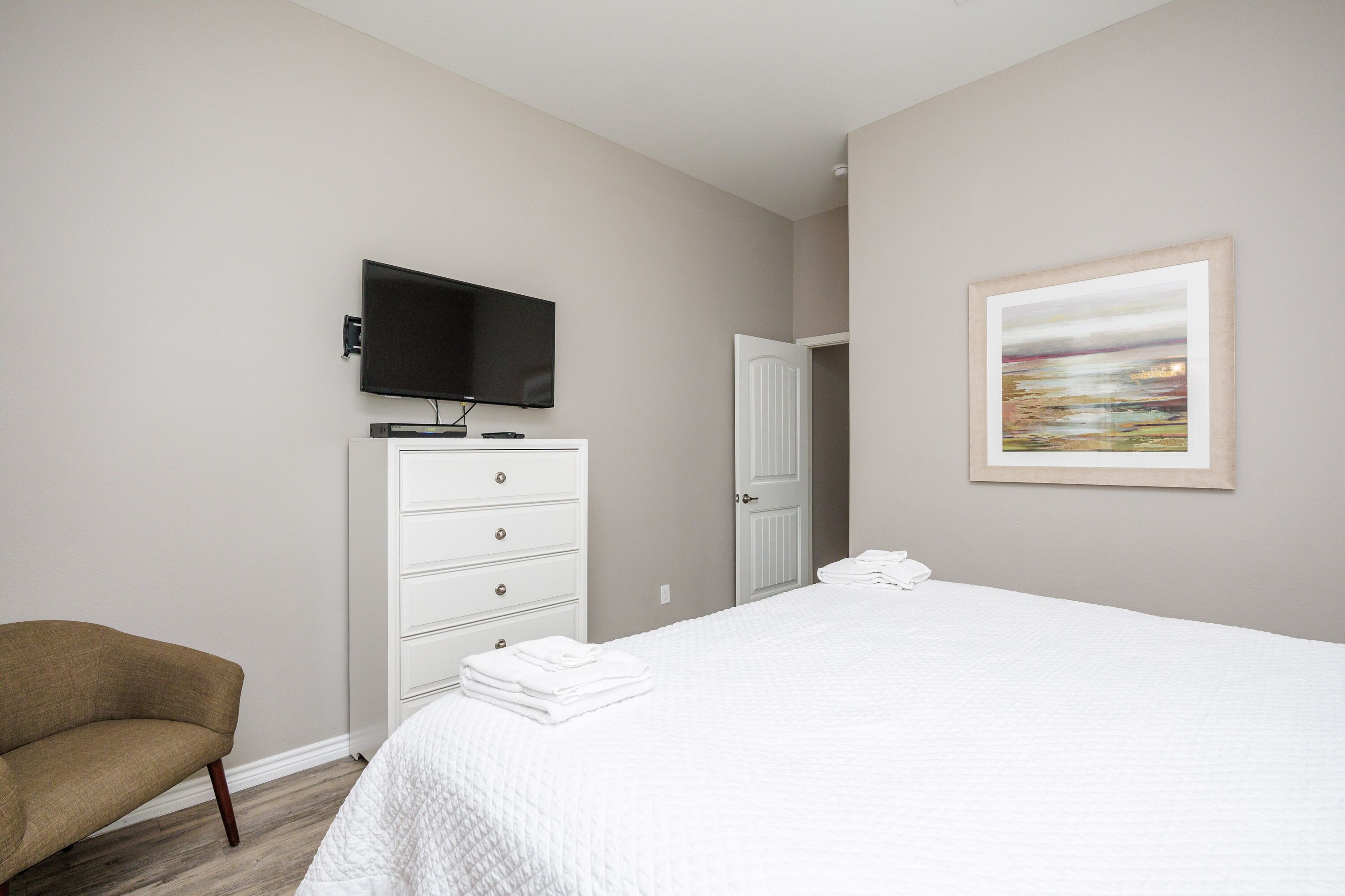 Une chambre d’hôtel avec un lit, une télévision, une commode blanche, une chaise et un tableau au mur.