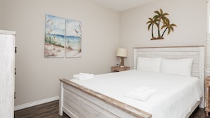A bedroom with a wooden bed, white bedding, a nightstand with a lamp, and two beach-themed wall art pieces.