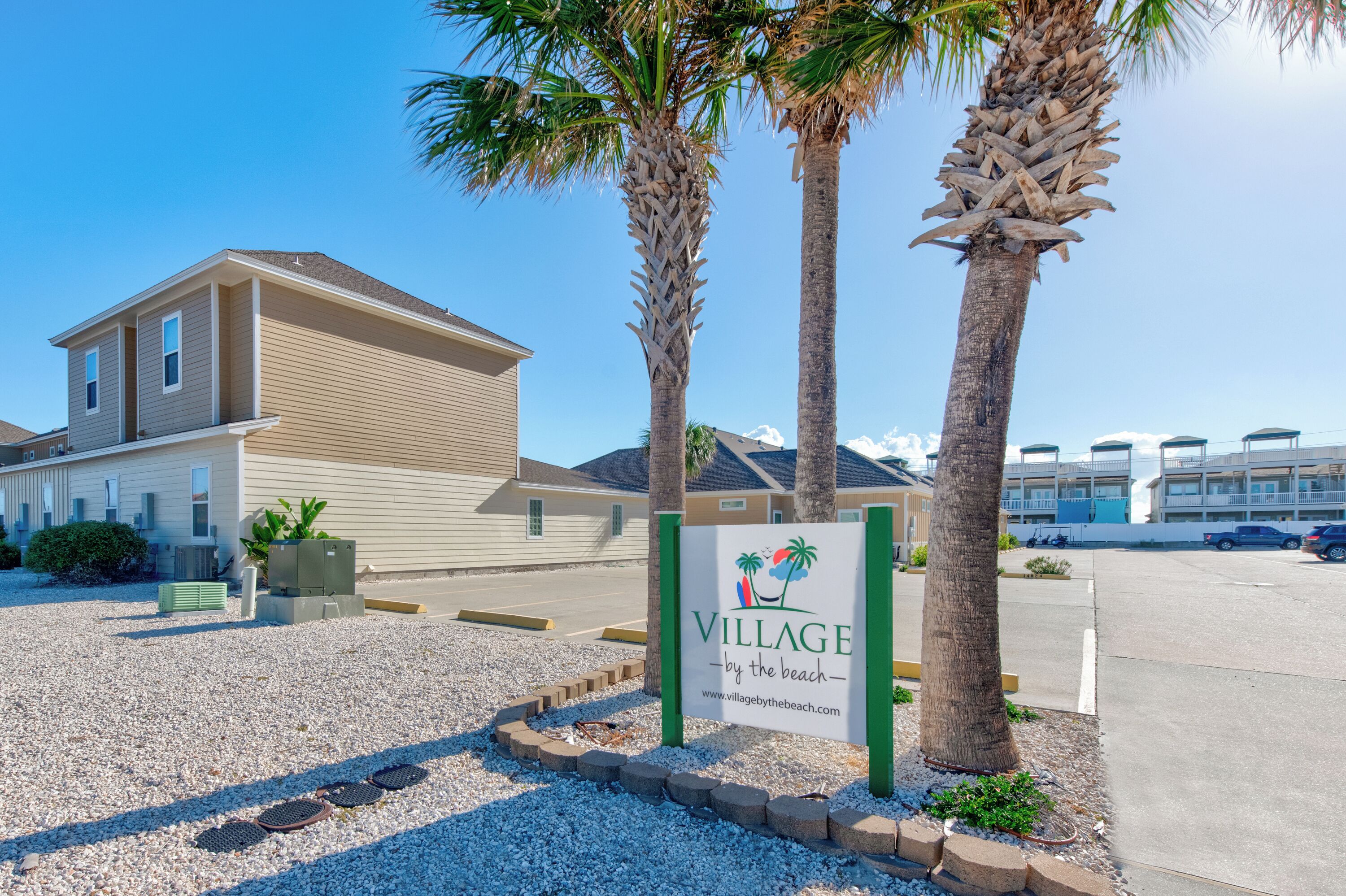 Village by the Beach entrance with coastal palm trees and vacation rental buildings in tropical setting.