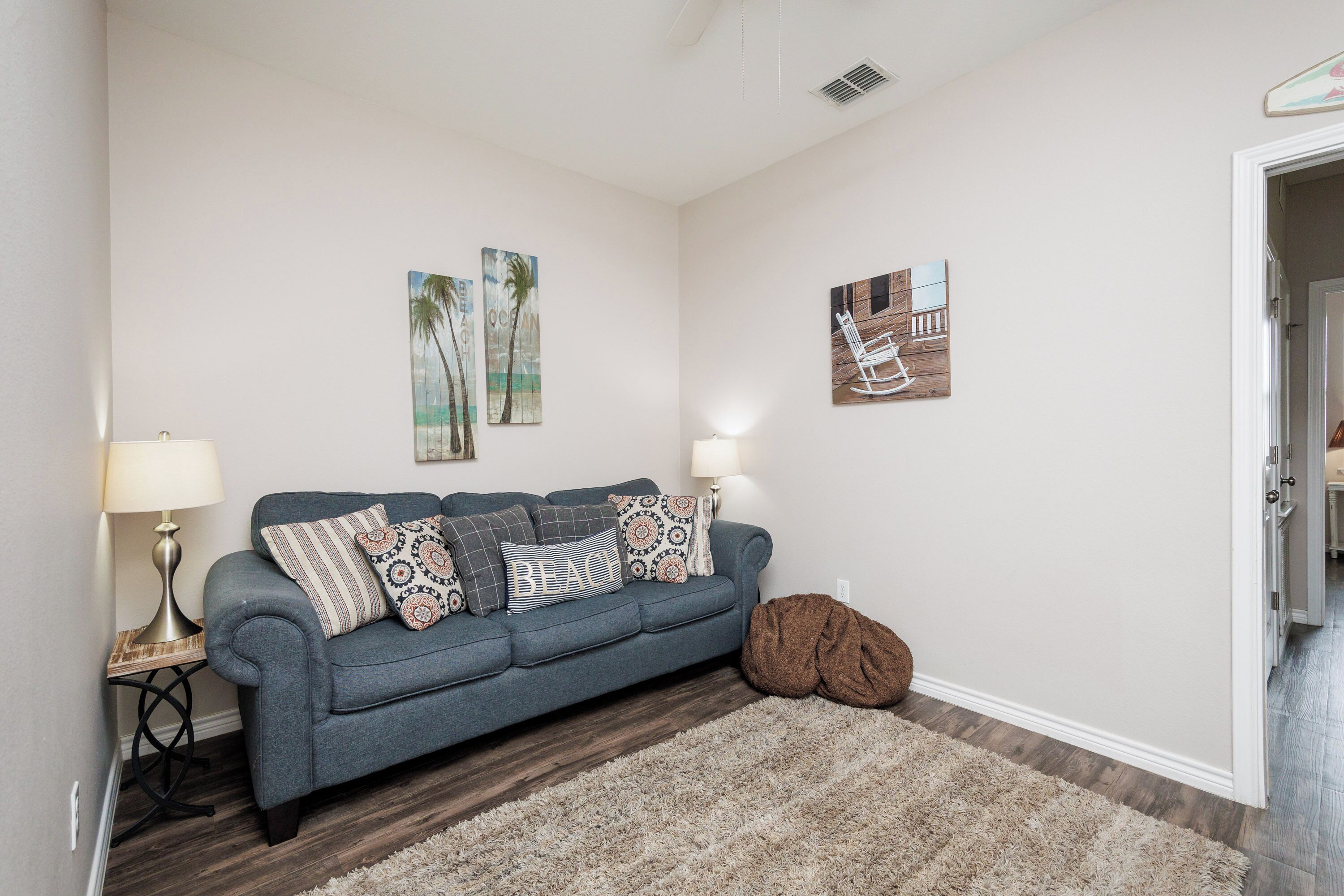 A living room with a blue sofa, two lamps, a brown rug, and wall art depicting palm trees and a rocking chair.