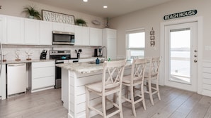 A kitchen with white cabinets, a central island with bar stools, a microwave, and a sign that reads "SANDY FEET WELCOME". - Pet-Friendly Retreat | 2 Resort Pools, Pickleball & Docks (Corpus Christi)