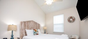 A bedroom with a bed, two bedside tables with lamps, a ceiling fan, a window with blinds, and a decorative wreath on the wall.