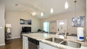A modern kitchen with a marble countertop, stainless steel sink, and a dishwasher. There is a ceiling fan, pendant lights, and a flat-screen TV. - Your Perfect Escape | Heated Pools, Pickleball & Resort Amenities (Corpus Christi)