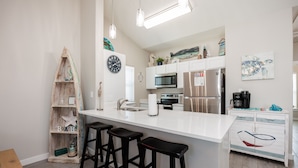 A modern kitchen with a large island, bar stools, a wooden shelf with decorative items, a clock, and a turtle painting. - Luxury Townhome | Private Patio, 2 Heated Pools & Amenities (Corpus Christi)